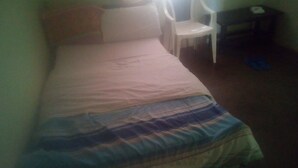 A small, single-bed room with a plain bedspread, a white chair, and a small desk with a chair.