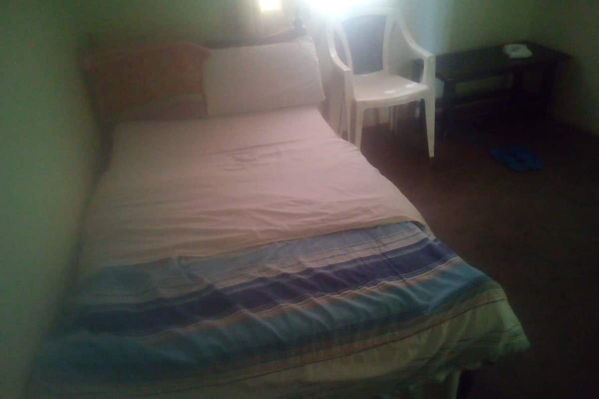 A small, single-bed room with a plain bedspread, a white chair, and a small desk with a chair.