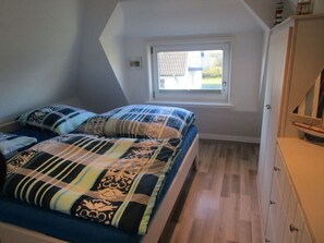A small, well-lit room with a sloped ceiling, a single bed with patterned bedding, a window with a view of a house, and a wooden cabinet.