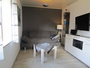 A compact living space with a grey sofa, a small table with a blanket, a lamp, a TV, and a bookshelf.