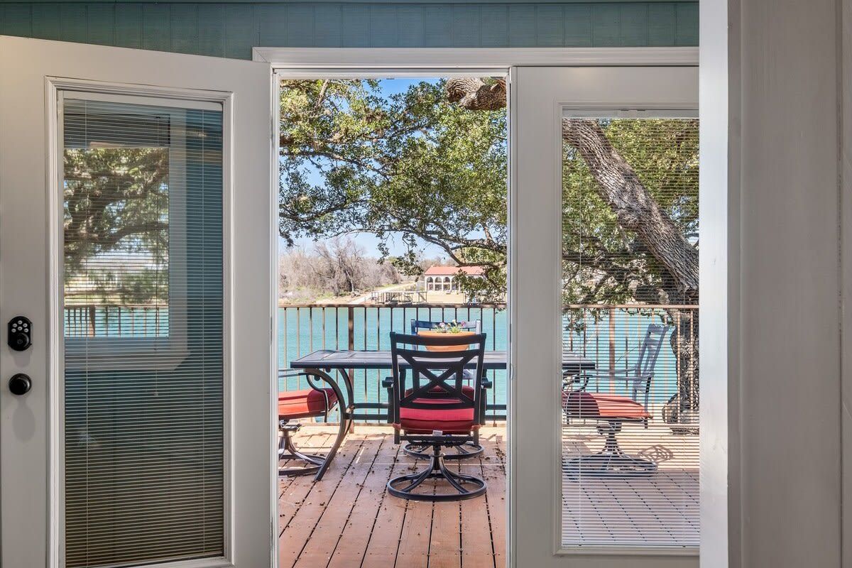 A view through a door to an outdoor deck with a table and chairs, a pool, and a fence.