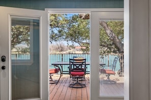 On aperçoit, à travers une porte, une terrasse extérieure avec une table et des chaises, une piscine et une clôture.