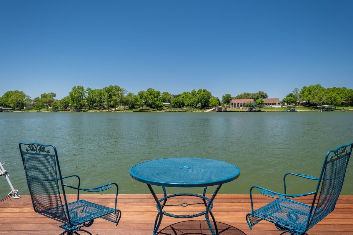 Eine Holzterrasse mit zwei Metallstühlen und einem Tisch, mit Blick auf einen stillen See unter blauem Himmel.