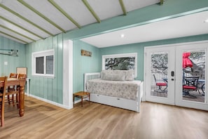 A bedroom with a bed, a small table, a window, and a door leading to an outdoor area.