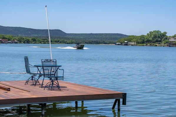 Un muelle de madera con una mesa y sillas se extiende sobre un lago tranquilo, donde se divisa un barco a lo lejos y un paisaje montañoso al fondo.