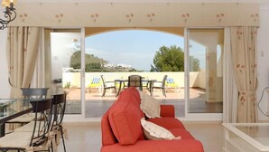 A living room with a red sofa, a glass dining table, and chairs. There is a view of a patio with a table and chairs outside.