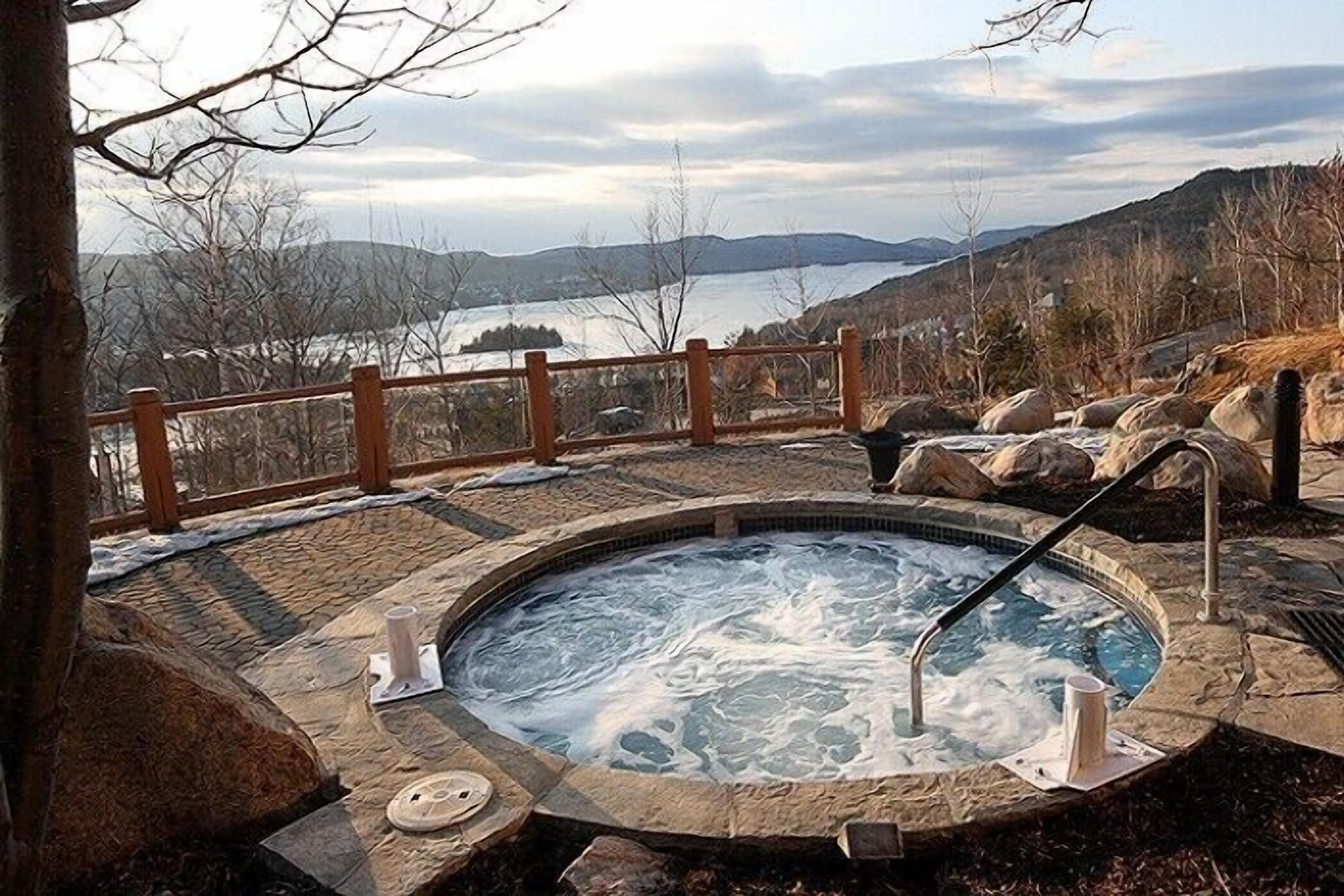 A hot tub surrounded by stone tiles, with a wooden fence and a view of a lake and mountains in the background.