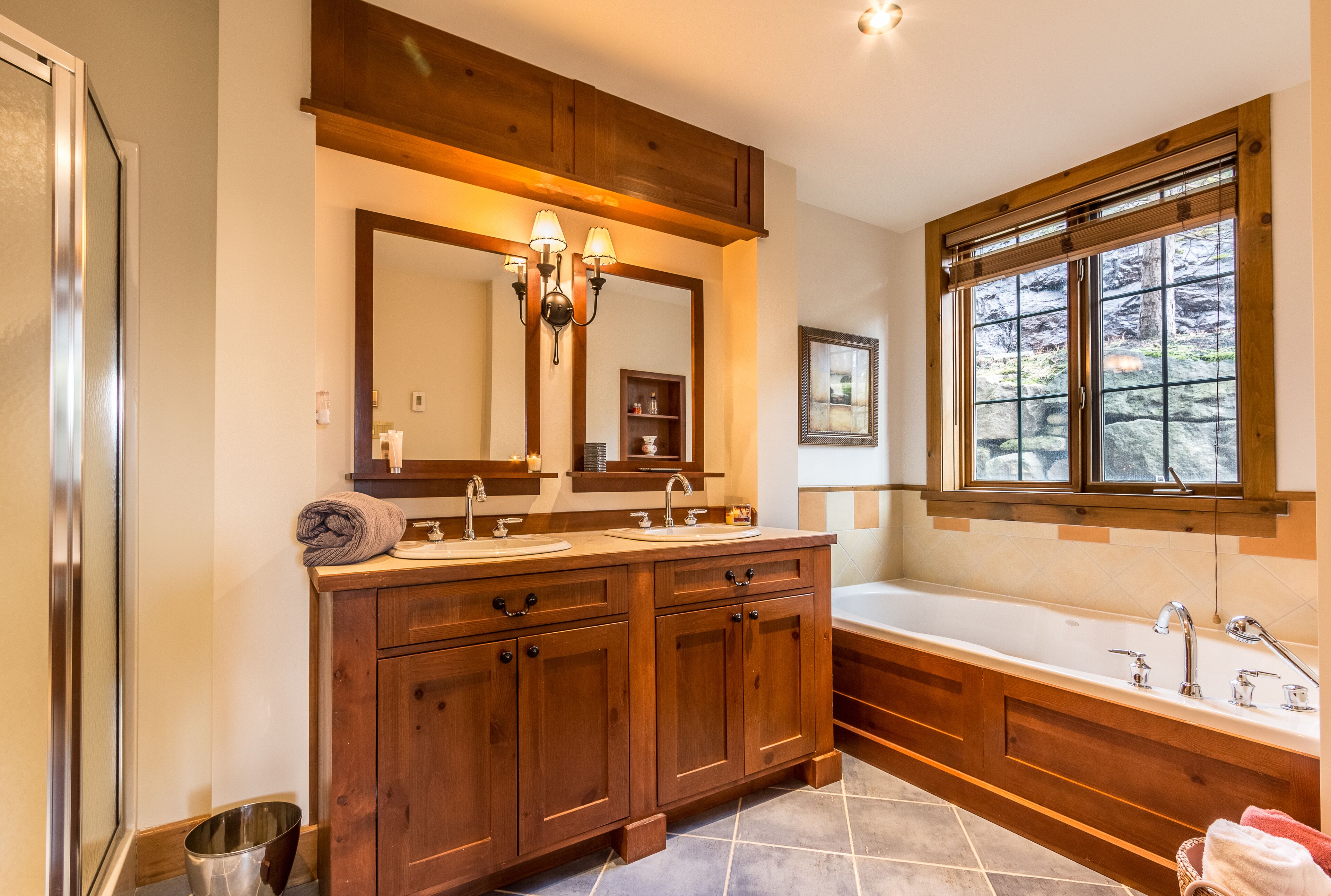 A bathroom with wooden cabinetry, a large mirror, a double sink vanity, a bathtub, and a window with a view of trees.
