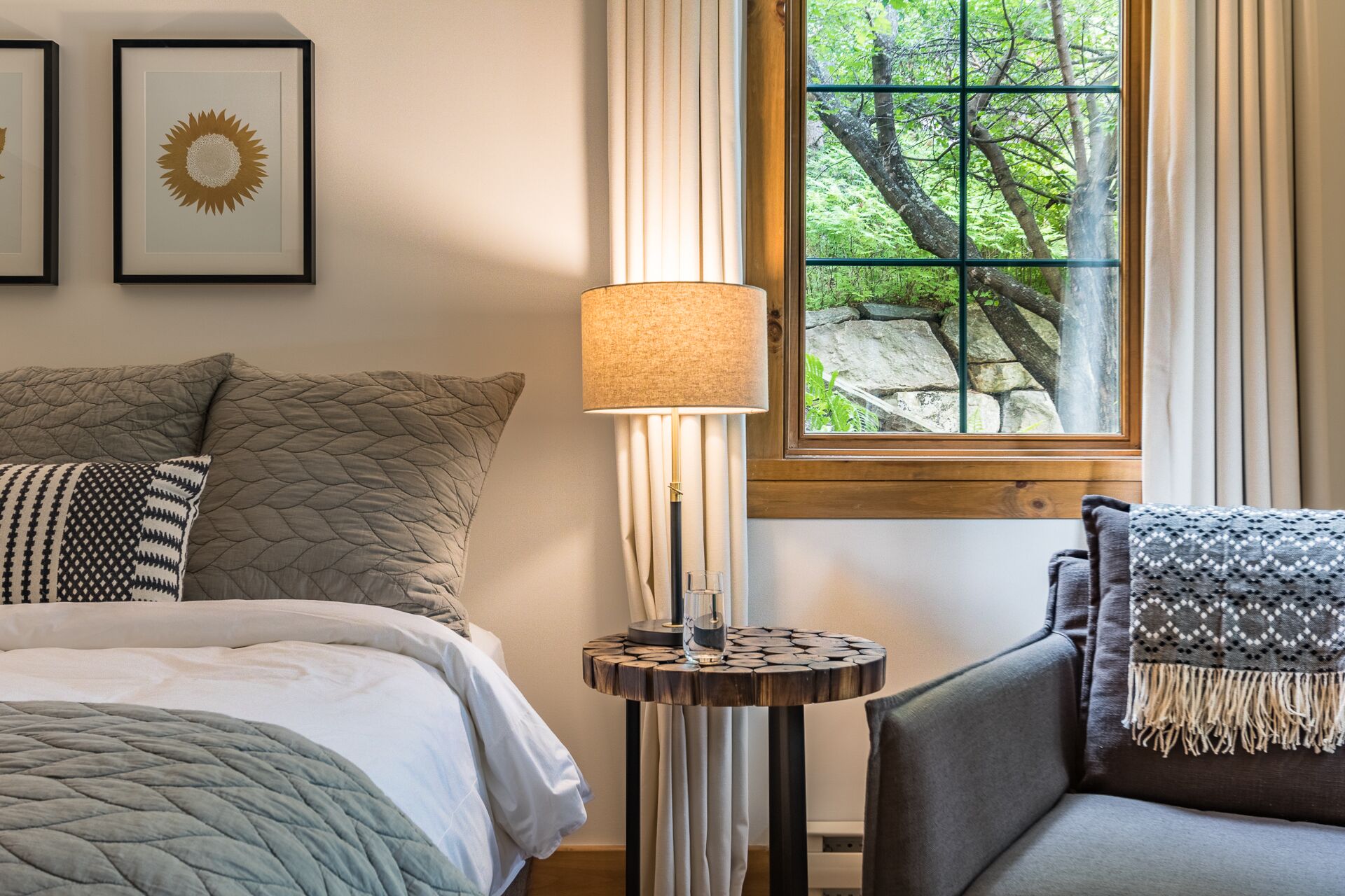 A neatly arranged bedroom with a bed, a bedside table with a lamp, a chair, and a window with a view of trees.