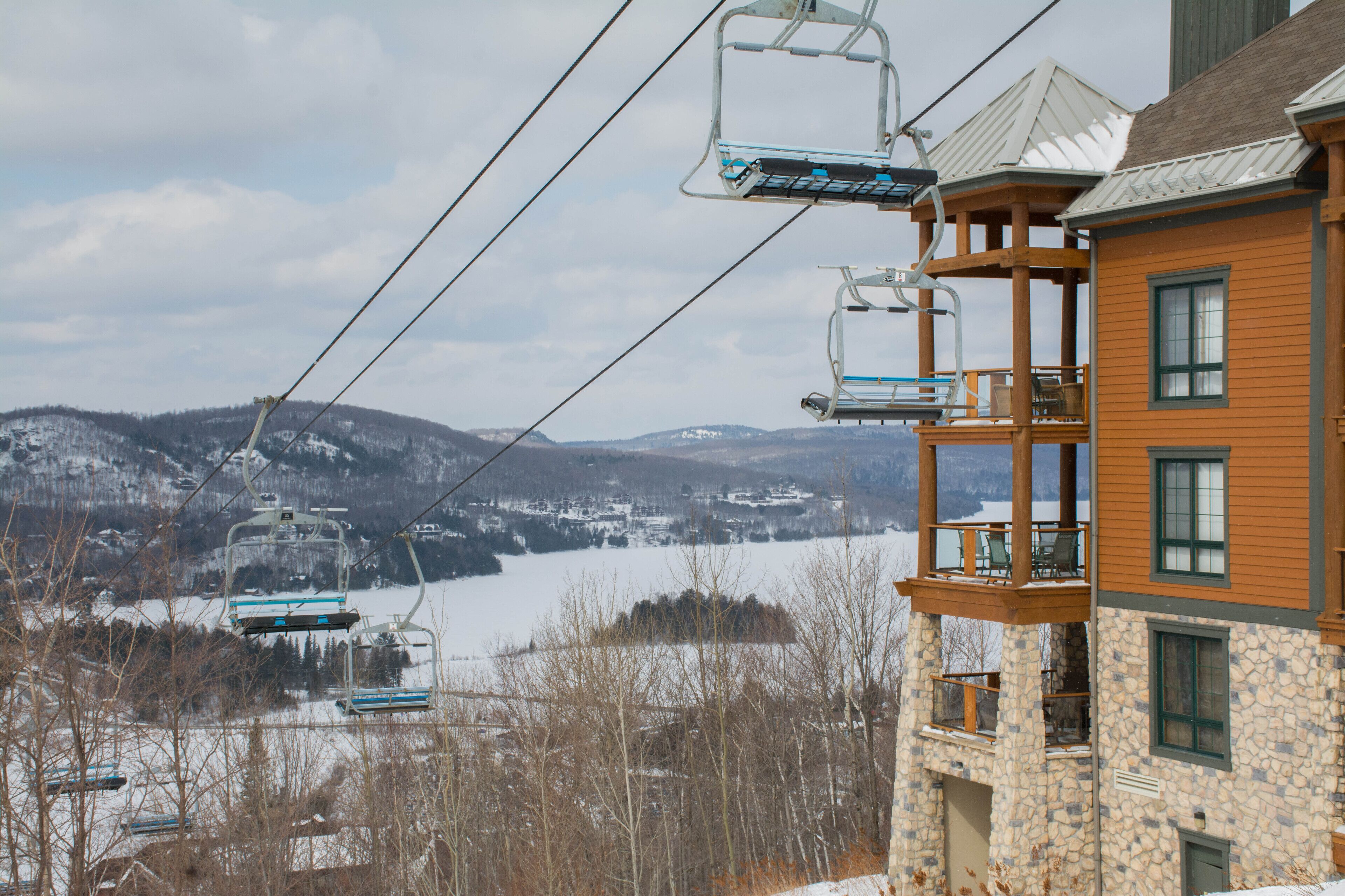 A ski lift with chairs, a stone building with a balcony, and a snowy landscape in the background.