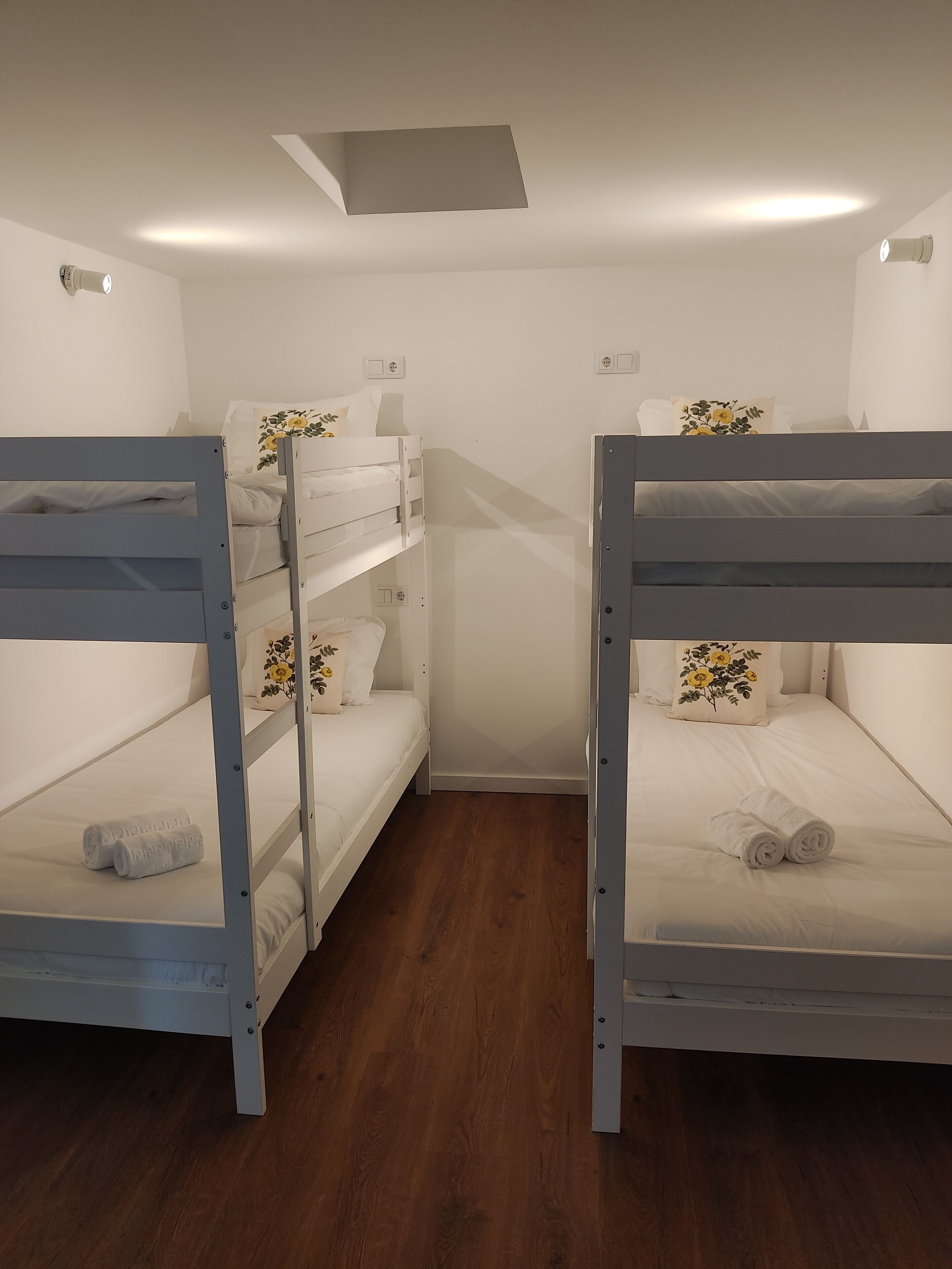 A room with two bunk beds, wooden flooring, white walls, and a small skylight.