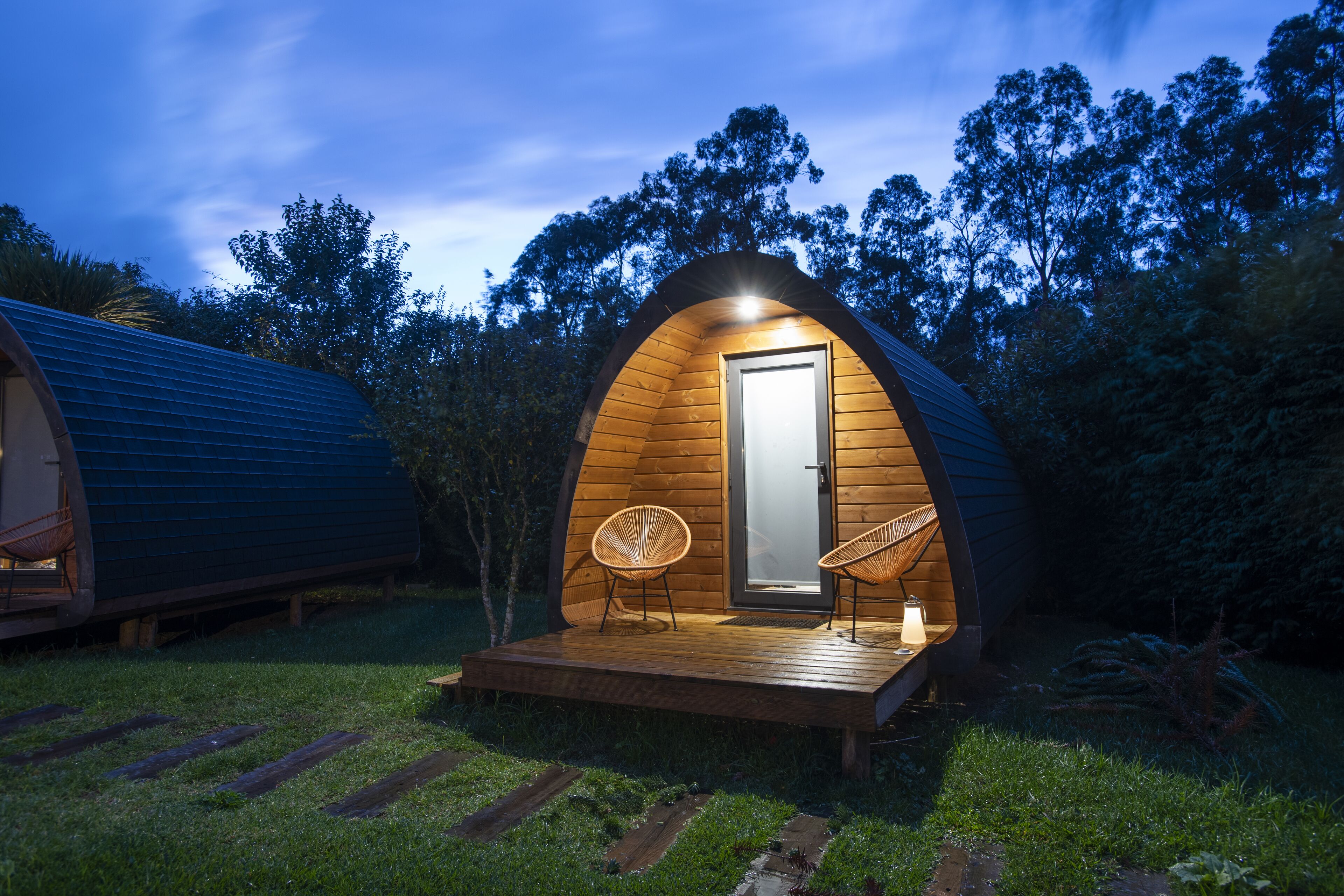 Ein gemütliches Holz-Viereckhaus mit Veranda, zwei Korbstühle und einem kleinen Tisch mit Lampe.