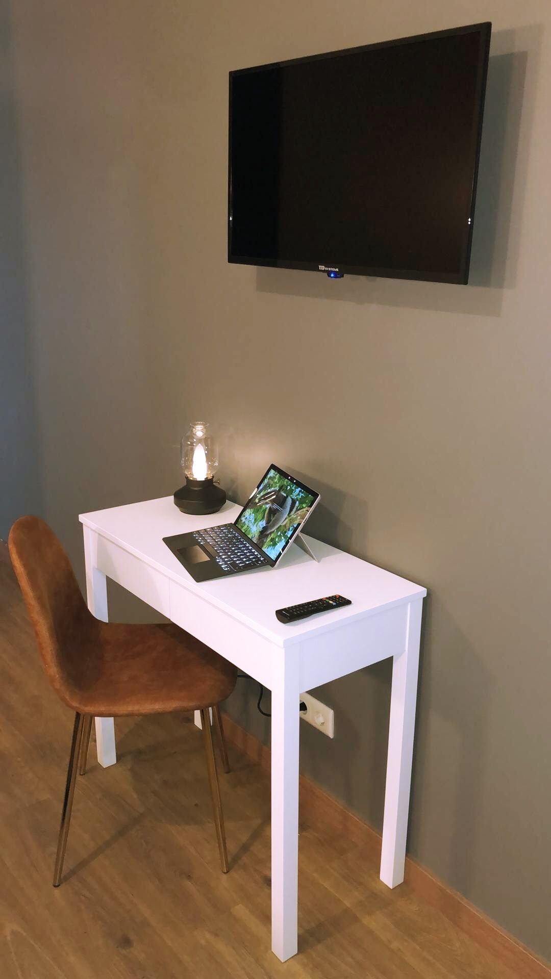 A small room with a white desk, a laptop, a remote control, and a TV mounted on the wall.