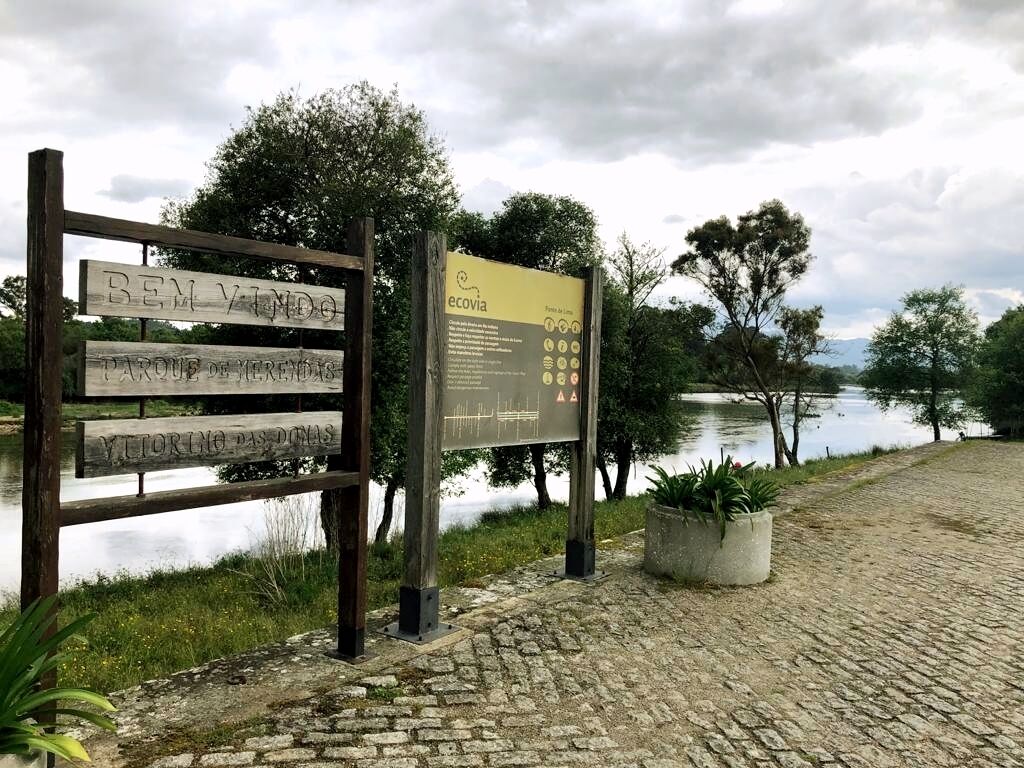 A wooden sign with 'Bem Vindo' and 'Parque de Janelas' text, a placid lake, and a paved walkway.