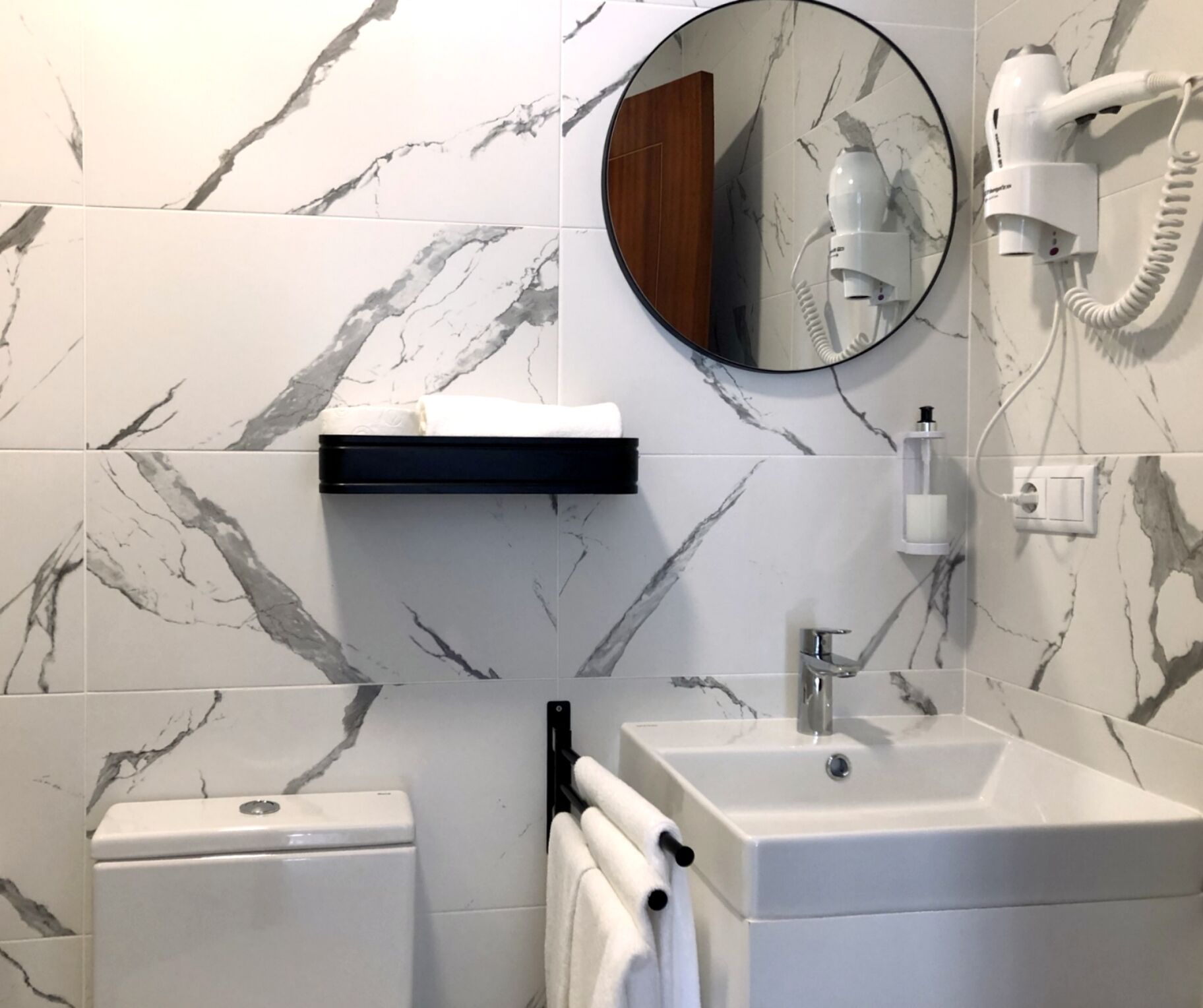 Um banheiro com painel de parede de mármore, um espelho redondo, uma pia branca, um vaso sanitário e um porta-toalhas.