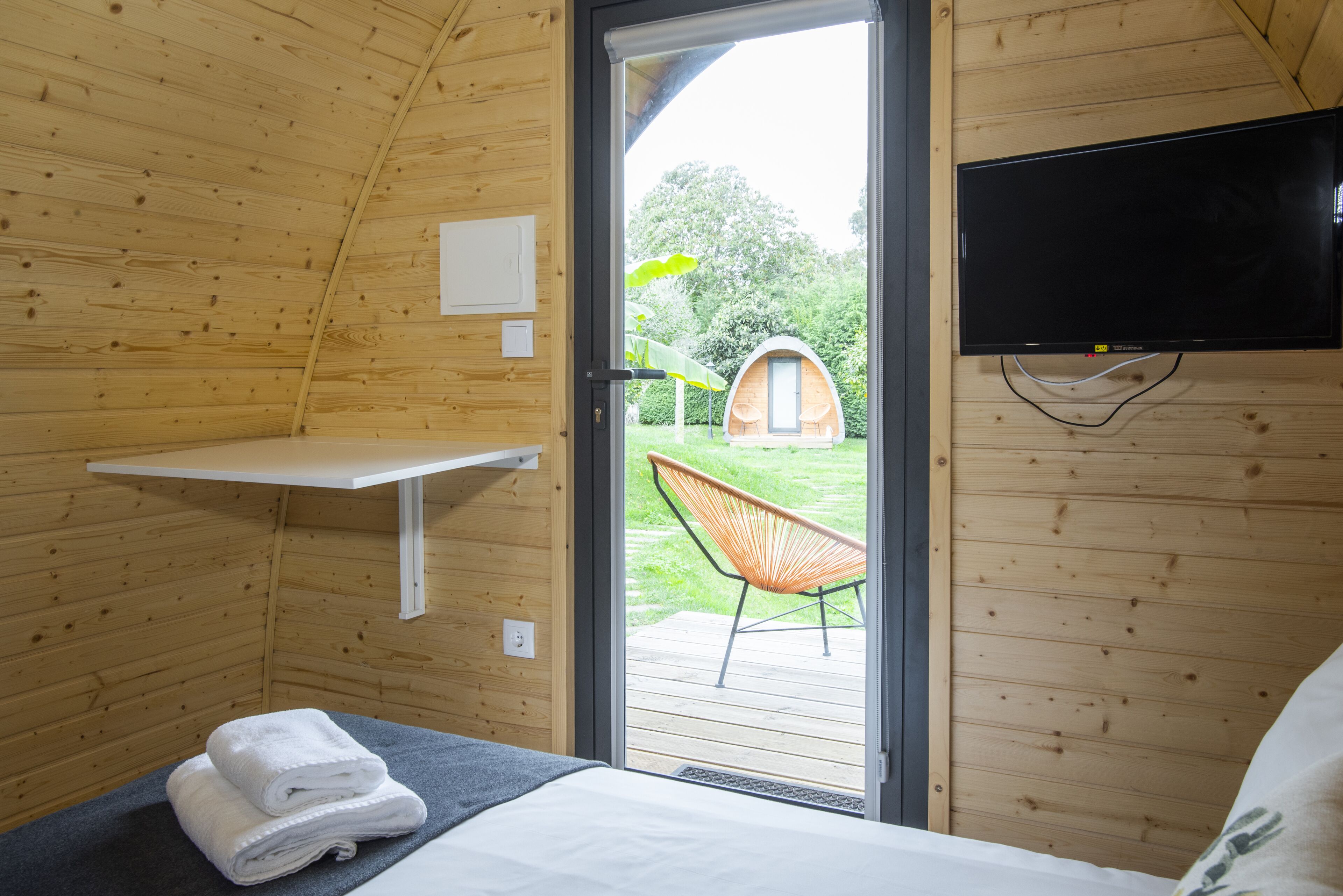 A cozy wooden room with a bed, a TV, and a door leading to an outdoor area with a chair and a small structure.