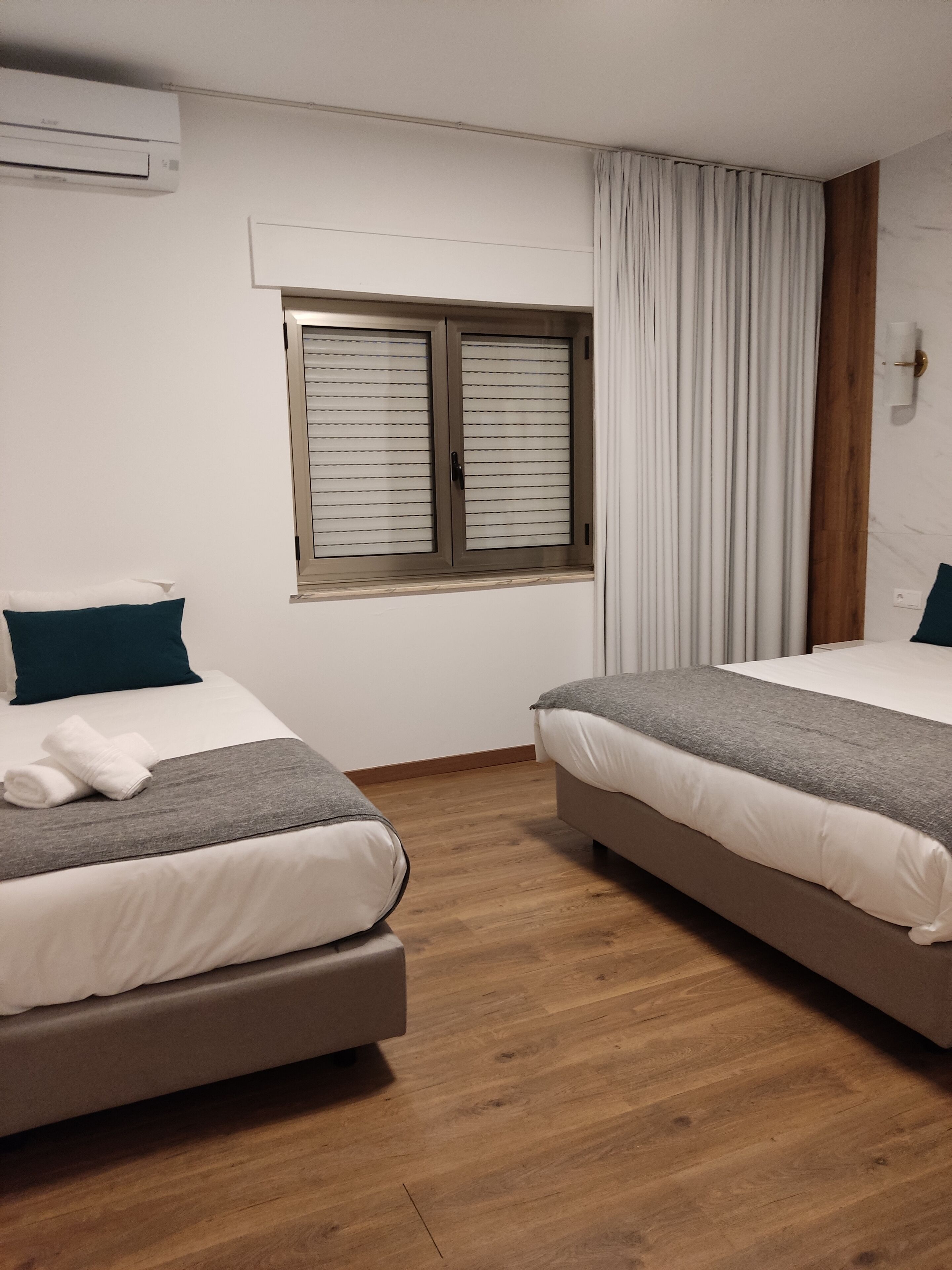 Une chambre d’hôtel avec deux lits, une fenêtre avec des stores, un plancher en bois et un climatiseur fixé au mur.