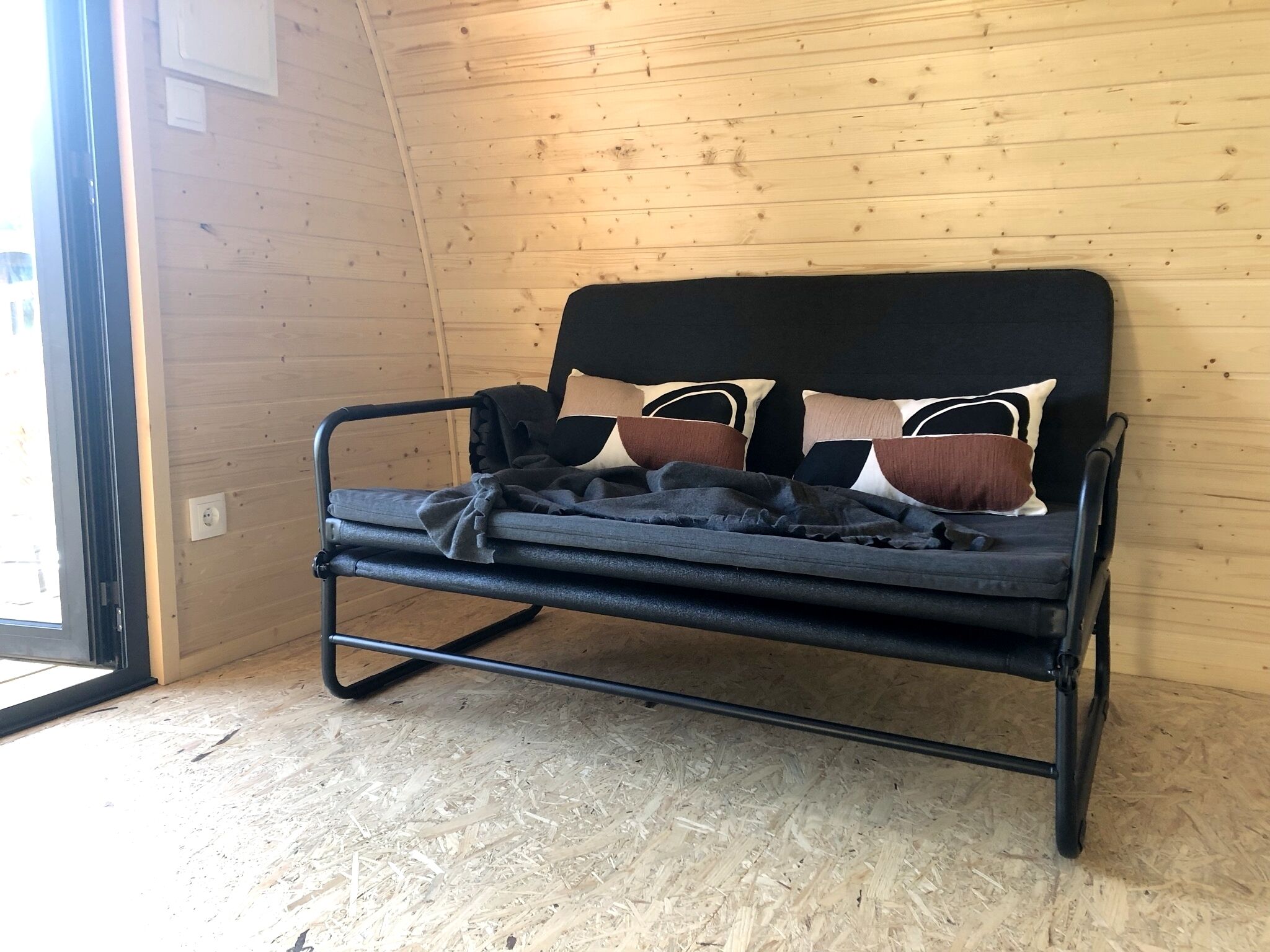 A metal-framed bed with a dark mattress and multiple pillows against a wooden wall.
