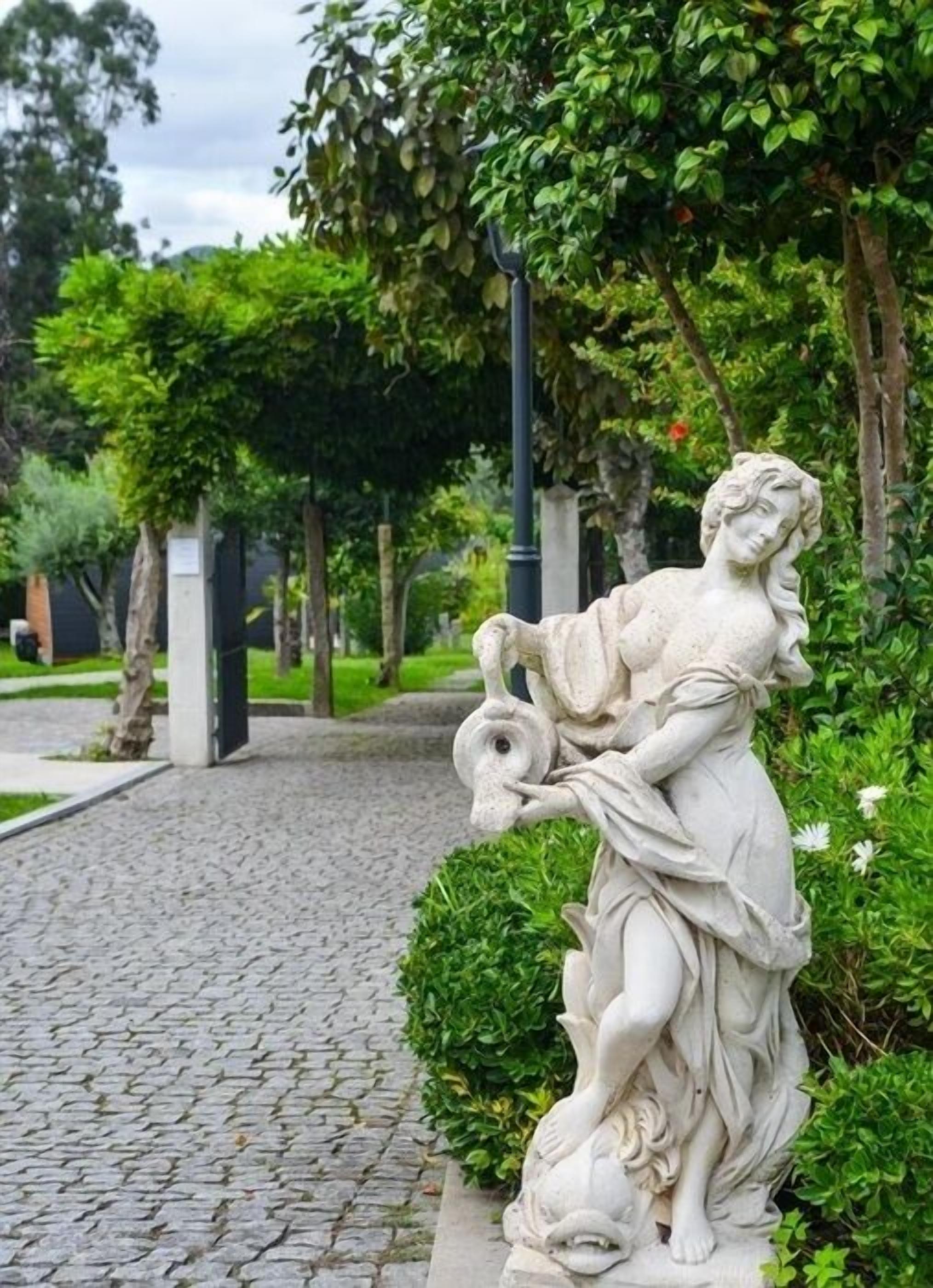 A stone statue of a woman holding a fish, located on a cobblestone path surrounded by greenery.