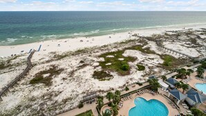 Eine Vogelperspektive zeigt einen Strand mit Poolbereich, einer Pier und einem weiteren Strandabschnitt mit Sonnenschirmen und Menschen.