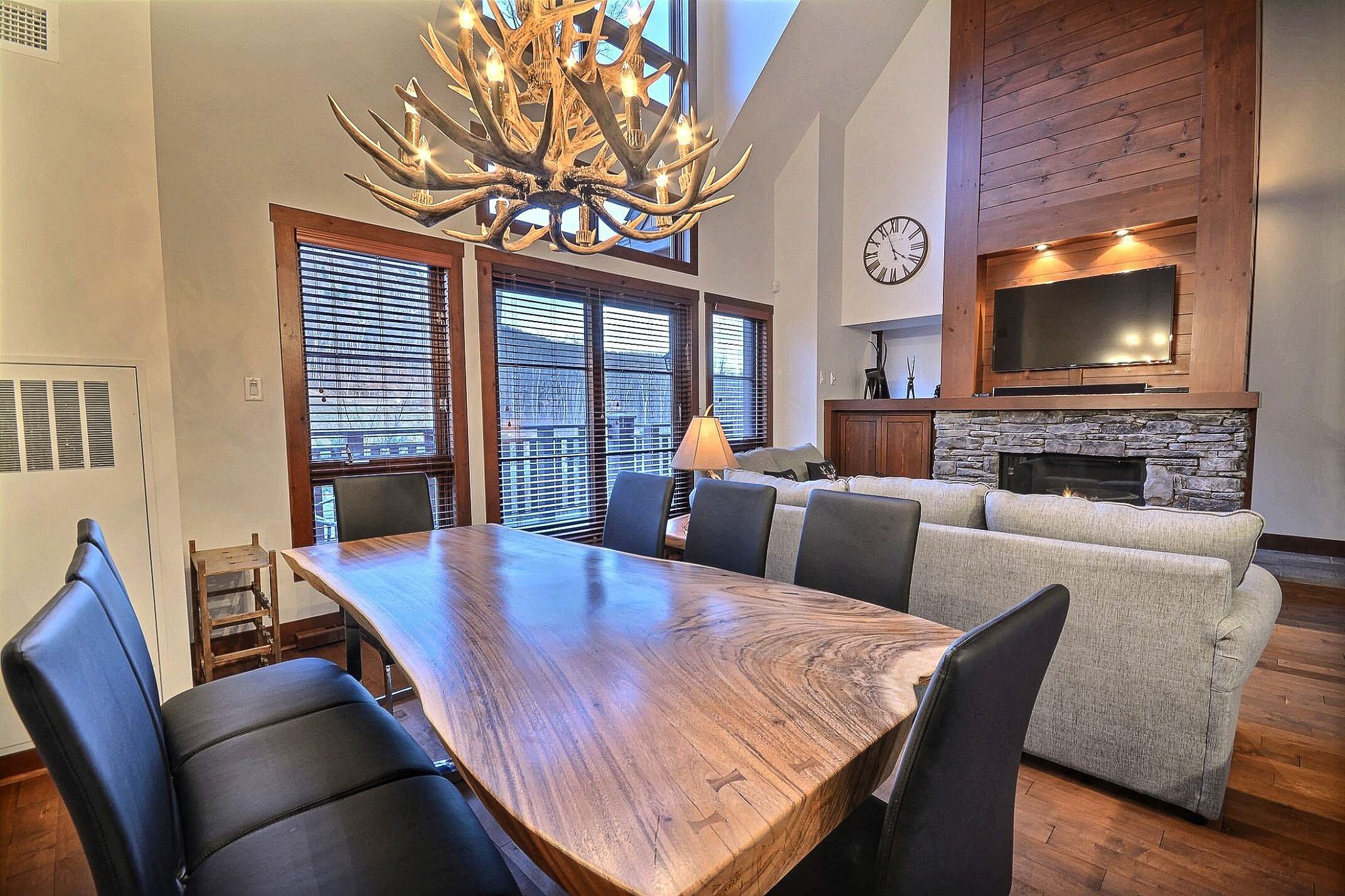 A dining area with a wooden table and chairs, a fireplace, and a large window with blinds.