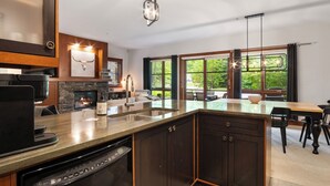 A modern kitchen with a built-in dishwasher, a stone fireplace, and a dining area with a large window overlooking greenery.