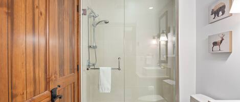 A bathroom with a glass shower enclosure, wooden door, and a white sink.