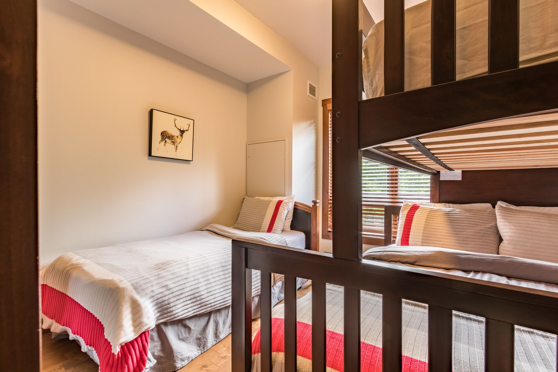 A bedroom with two beds, a wooden bunk bed, a red and white striped blanket, and a deer painting on the wall.