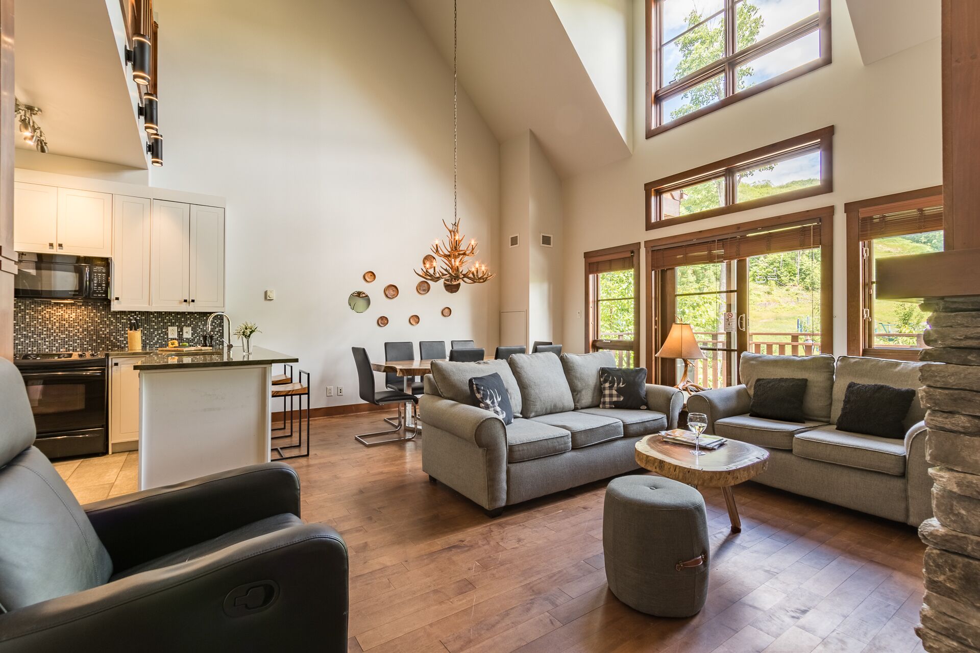 A spacious living area with a large sofa, a coffee table, and a dining table set for six. There is a kitchen with modern appliances and a chandelier hanging from the ceiling.