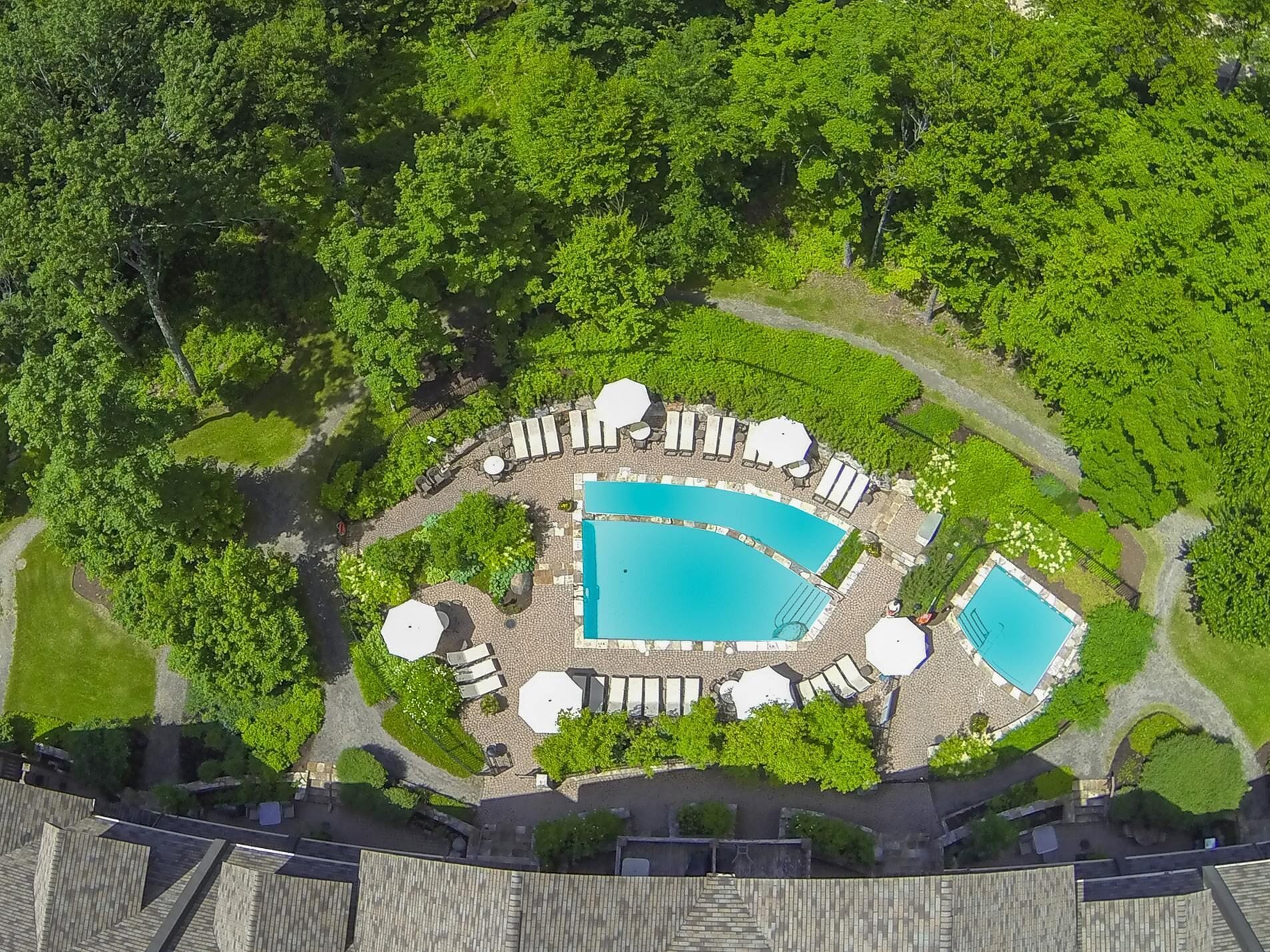 An aerial view of a resort with two swimming pools surrounded by lounge chairs and umbrellas, set in a lush green landscape.