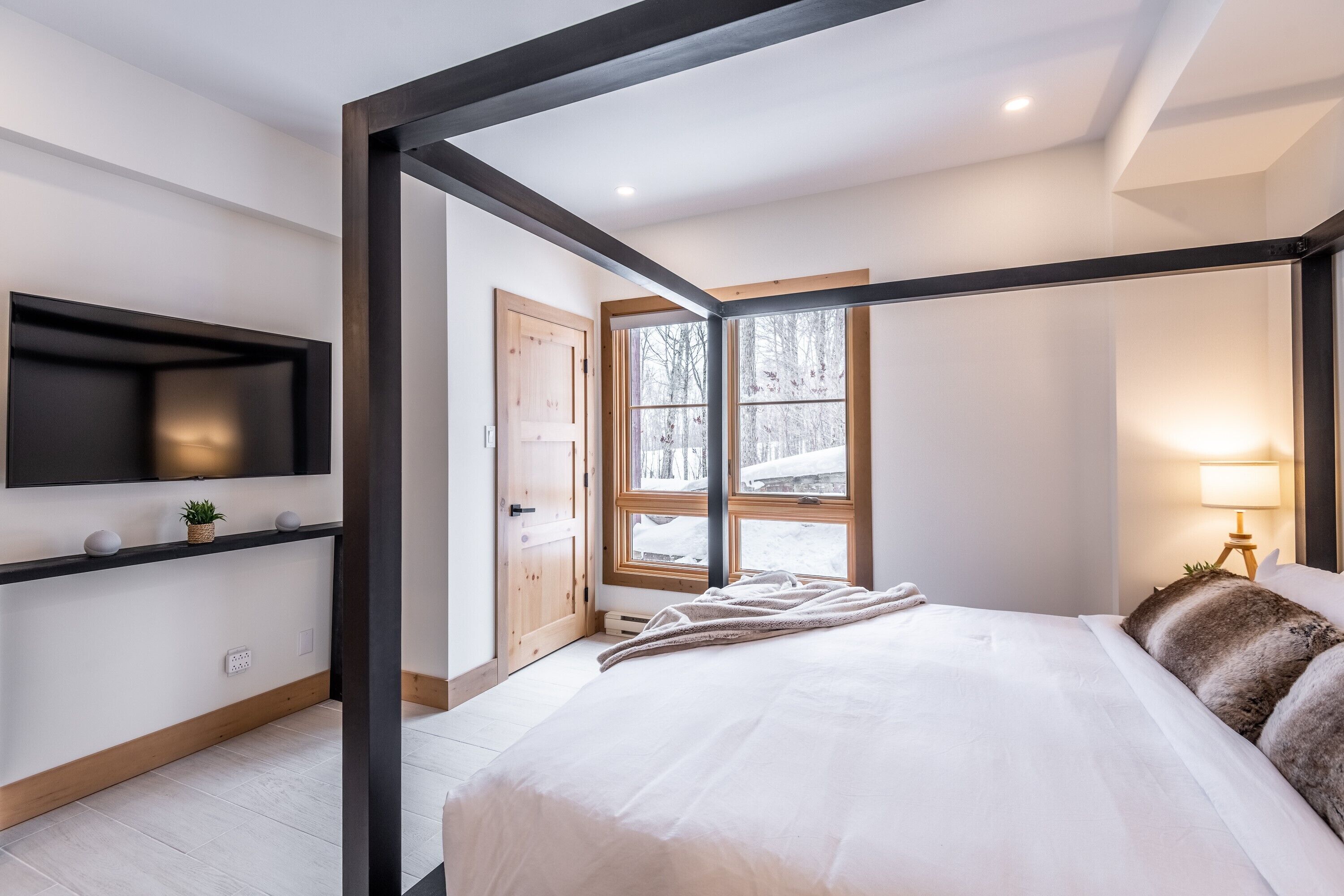 Un dormitorio moderno con una cama grande, un televisor montado en la pared y una ventana con vista a árboles.