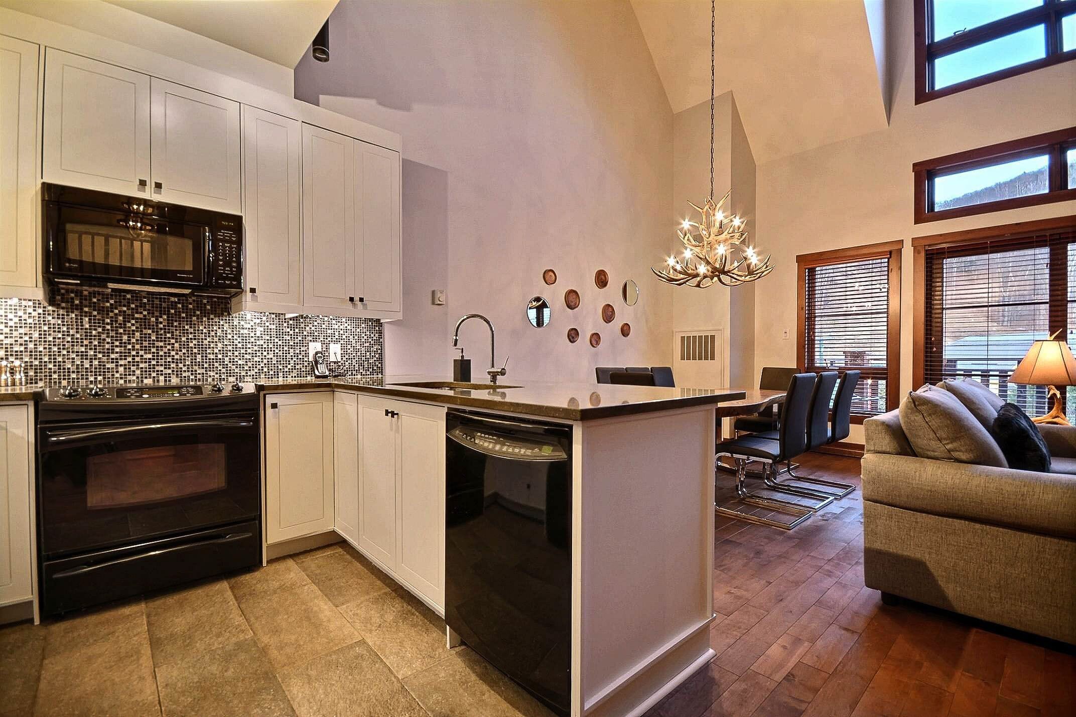 A modern kitchen with white cabinets, a black microwave, and a dishwasher. There is a dining area with a sofa and a chandelier.