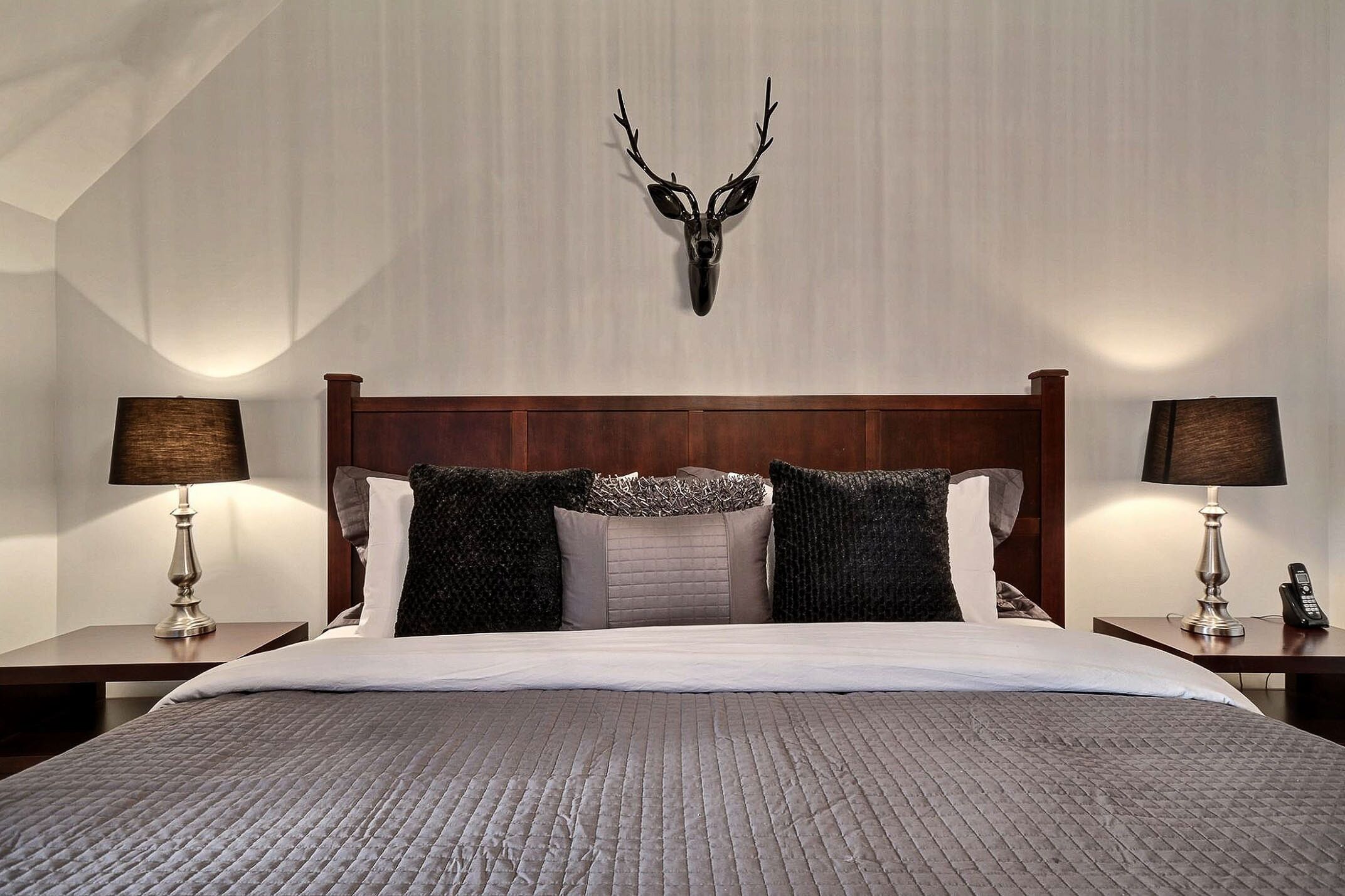 A neatly made bed with a headboard, two bedside lamps, and a mounted deer head on the wall.