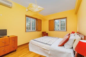 Quarto com cama, cómoda de madeira, televisão e duas janelas.
