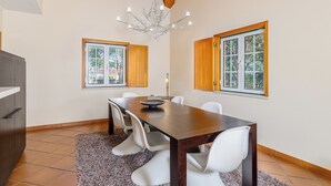 A dining area with a wooden table, white chairs, a chandelier, and windows with wooden frames. - Villa 97 with private pool by Trip2Portugal (Alcácer do Sal)
