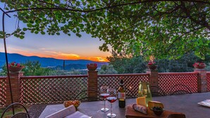 An outdoor dining area with a view of mountains, a sunset, and a table set with wine glasses, cheese, and olives. - Poggio Medei 10, Emma Villas (Città della Pieve)