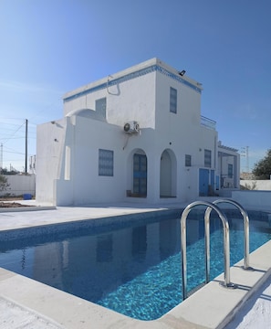 A modern building with a swimming pool, a covered outdoor area, and a clear blue sky.