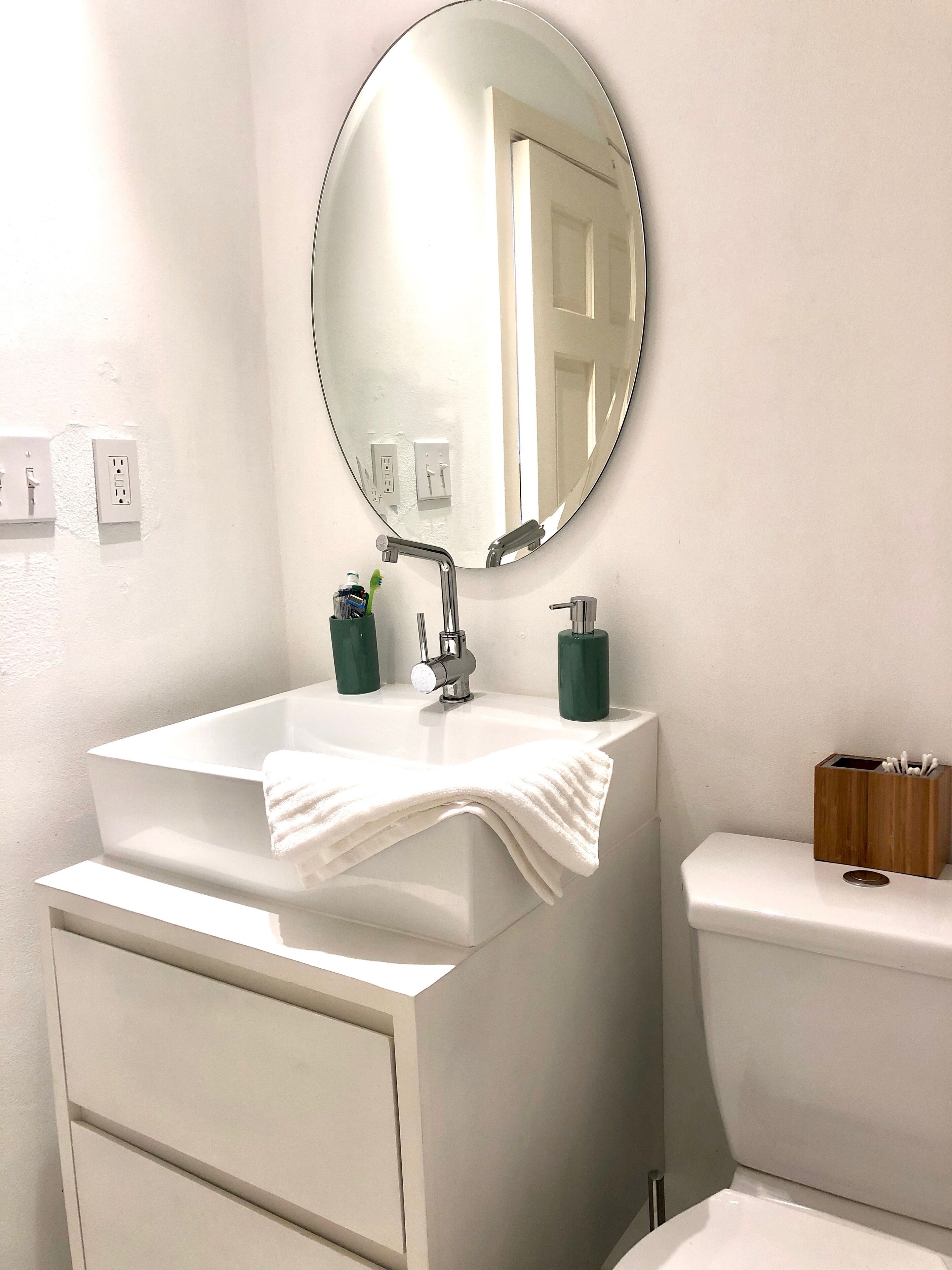 Une salle de bain avec un lavabo blanc, un miroir et des toilettes. On trouve deux récipients verts sur le lavabo et une serviette blanche posée sur le lavabo.