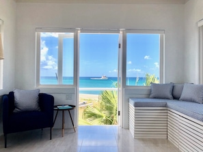 A room with a large sliding glass door leading to a balcony with a view of the ocean and a boat. There is a dark blue sofa with patterned cushions and a small round table with a green cushion.