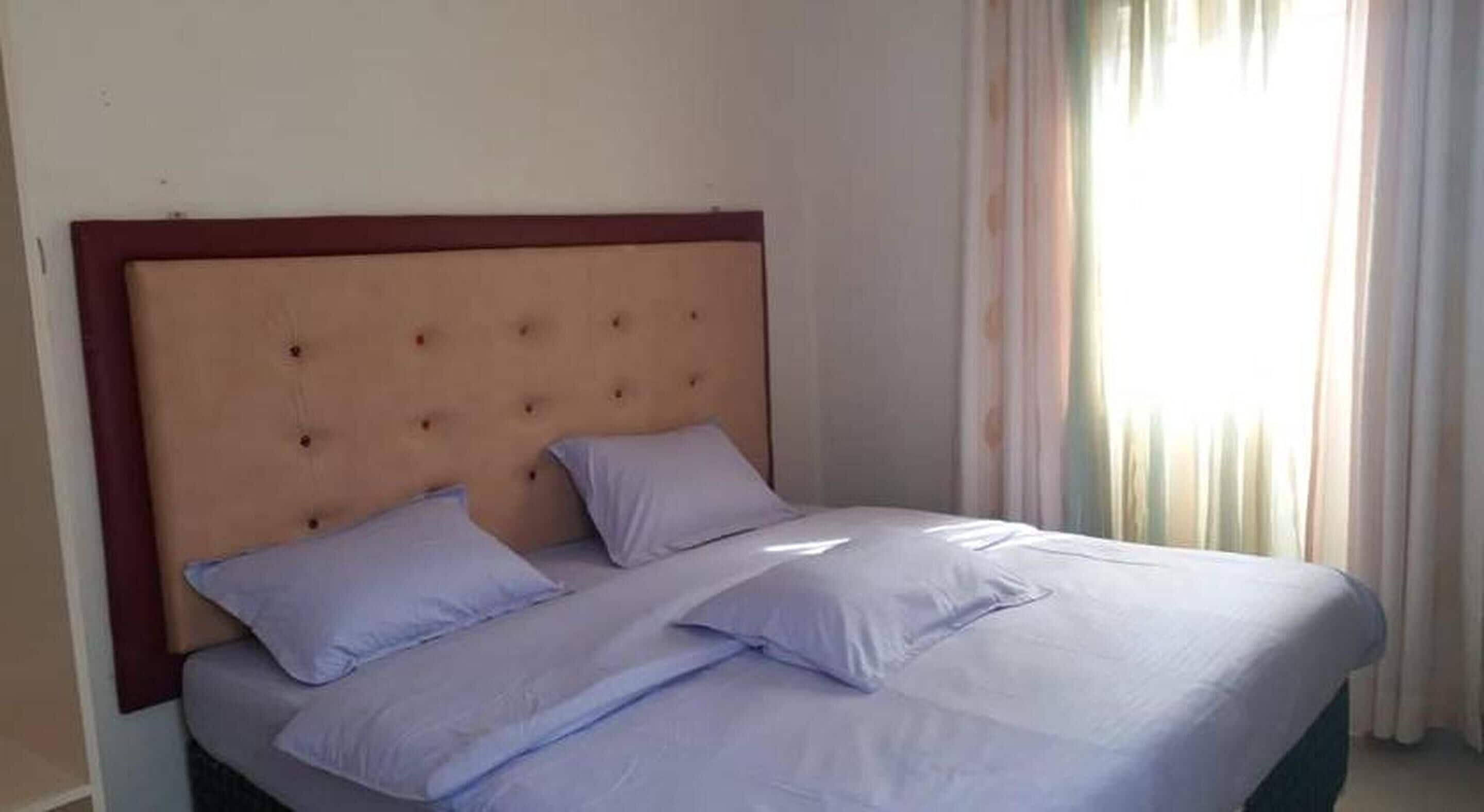 A bed with a wooden headboard and two pillows, a window with curtains, and a plain wall.