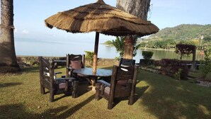 An outdoor dining area with a table and chairs under a thatched umbrella, overlooking a lake and hills. - Family Nest Kivu Beach (Nyayumba)