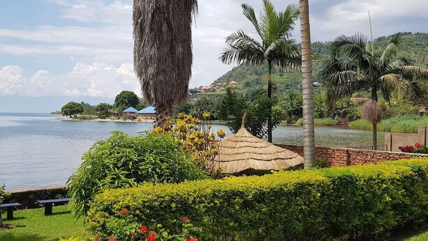 A serene lakeside garden with a thatched-roof gazebo, lush greenery, and a view of the lake and distant buildings. - Family Nest Kivu Beach (Nyayumba)