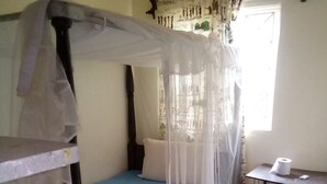 A single bed with a mosquito net, a small desk with a chair, and a window with curtains. - Paulice Guest House (Kisumu)