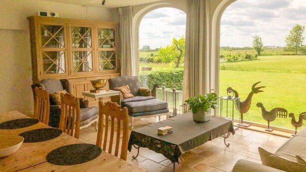 A spacious living area with a dining table, chairs, a sofa, and a cabinet. There are large windows offering a view of a green landscape. - Guest house beautiful historic square farm 🎯 in Ranst (Broechem Antwerp)