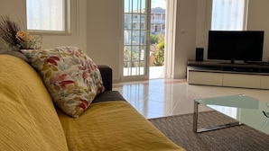 Ein Wohnzimmer mit einer Couch, einem Glastisch, einem Flachbildfernseher und einem großen Fenster mit Blick auf ein Gebäude und Grünflächen.