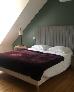 Ein Schlafzimmer mit schrägverdeckter Decke, einem Bett mit weißen Leinen, einer dunkelroten Tagesdecke, einer Nachttischlampe und einem Nachttisch.