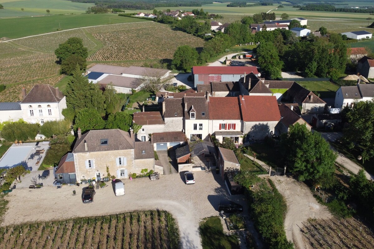 Eine Vogelperspektive auf ein ländliches Dorf mit traditionellen Steinhäusern, einem zentralen gepflasterten Platz mit parkenden Autos und umliegenden Ackerfeldern.