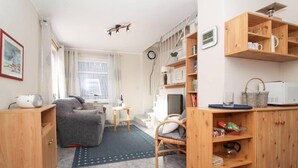 Un espacio habitable compacto con sofá, una pequeña mesa, una silla, una estantería y una zona de cocina con microondas.
