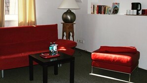 A living room with a red sofa, a red armchair, a small table, a lamp, a bookshelf with books, and a coffee machine. - Hotel Drei Kronen (Lippstadt)