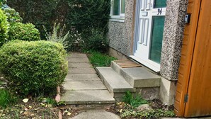 Un jardin résidentiel avec un chemin en béton menant à une maison dont la façade est en pierre et dont la porte porte le numéro 34.
